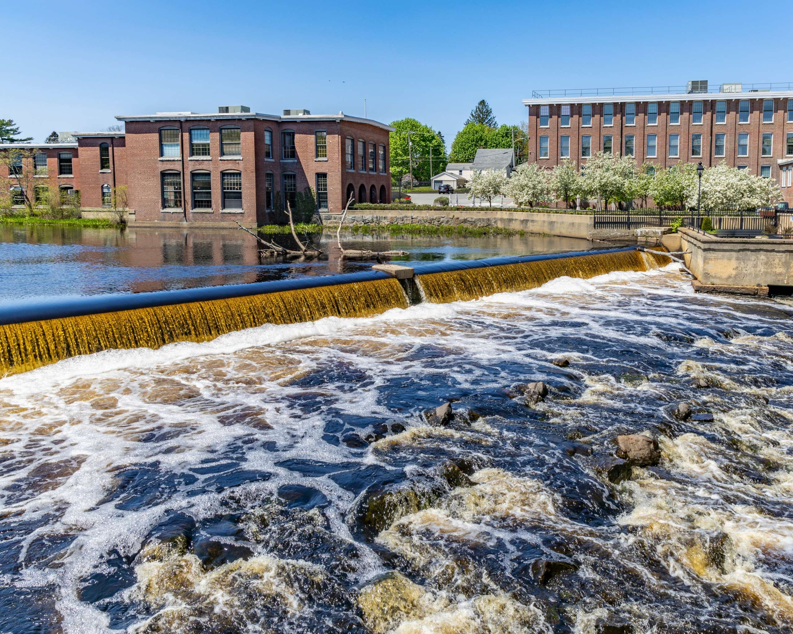 Water rushing over the Ipswich River dam in downtown Ipswich, with historic mill buildings lining the riverbank.
